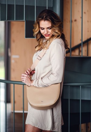 Beige vegan leather crossbody bag with a curved shape, adjustable patterned strap, and metallic logo detail. Smooth texture, minimalistic design.