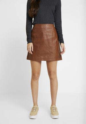 Woman wearing a brown leather mini skirt, dark long-sleeve top, and beige sneakers standing against a white background.
