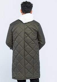 Lang, quiltet frakke i olivengrøn med diamant-syning, en hvid hætte og et slankt design. Materialet virker glat og let.