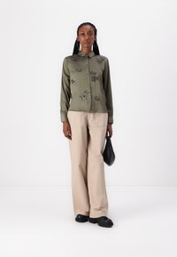 Olīvu zaļš satīna krekls ar ziedu izšuvumiem, apvienots ar bēšiem platajiem biksēm un melnām kurpēm, tur rokassomu melnā krāsā.