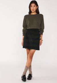 Maglione verde oliva con maniche raglan, abbinato a una mini gonna a quadri scuri con finiture a bottone e calze a maglia a trecce fino al ginocchio. Mocassini neri.