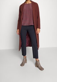 Modelka nosí burgundský kabát přes vzorovaný top, spárovaný s námořnickými kalhotami a taupe kotníkovými botami, stojící na jednolitém pozadí.