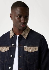 Chaqueta de mezclilla en azul oscuro con un cuello acolchado color beige que presenta un patrón de logotipos. Incluye botones plateados y dos bolsillos frontales.