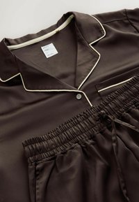 Brązowy satynowy zestaw piżamowy składający się z koszuli z kołnierzykiem z kremowym wykończeniem oraz kieszonką na piersi, do kompletu elastyczne spodenki.