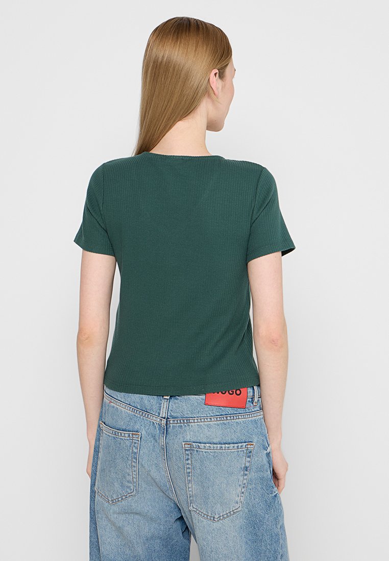 Grünes geripptes T-Shirt mit kurzen Ärmeln, rundem Halsausschnitt und cropped Passform. Kombiniert mit hellblauen High-Waist-Jeans mit einem roten Label.