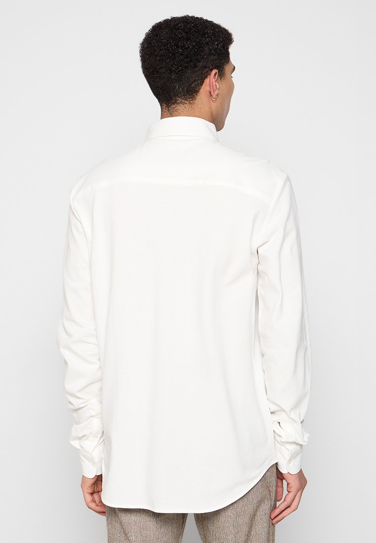 Biała, długosleeve koszula o luźnym kroju, z mankietami na guziki i gładką teksturą. Brak widocznych wzorów lub akcentów.