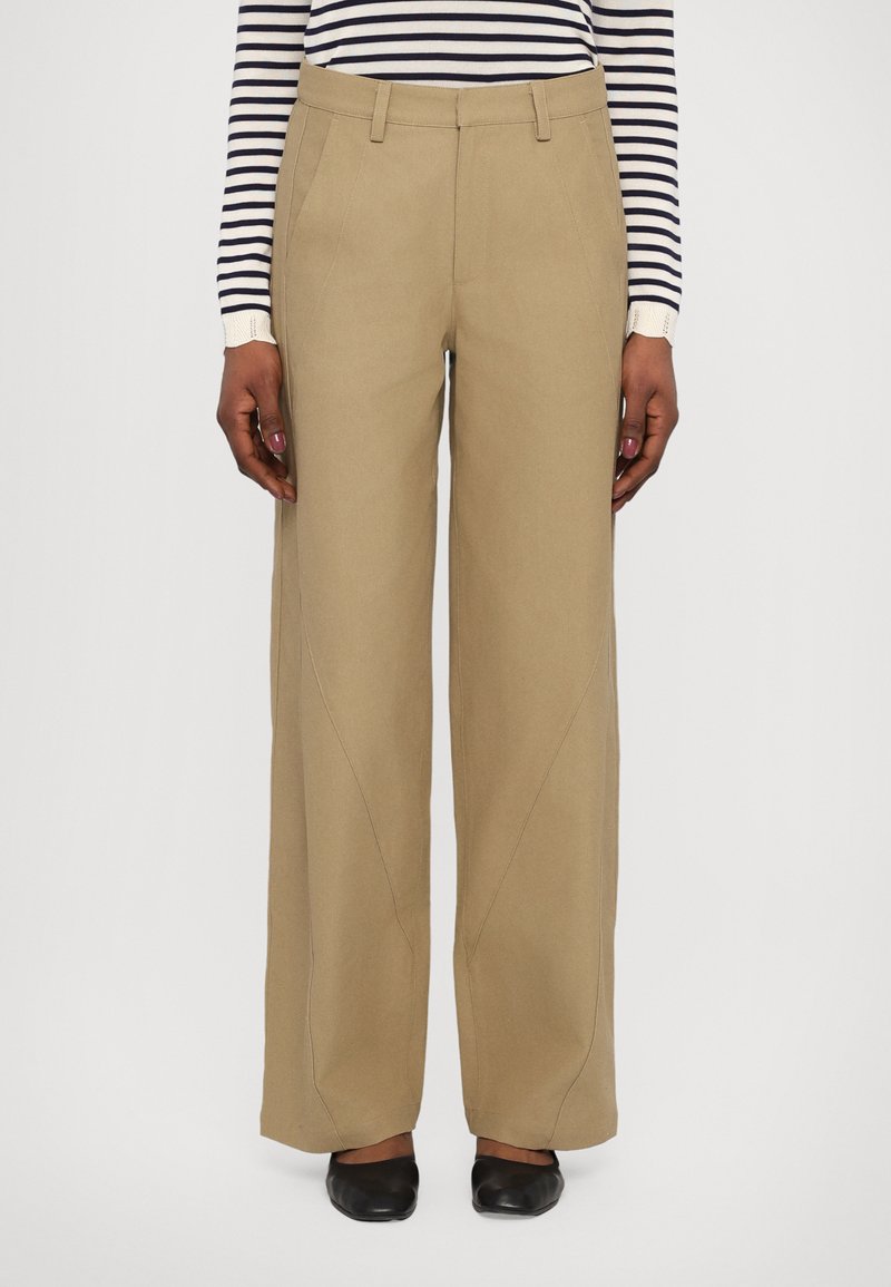 Pantaloni beige a gamba larga realizzati in tessuto resistente, con vita media, tasche frontali e dettagli delle cuciture sottili. Indossati con una maglietta a righe.