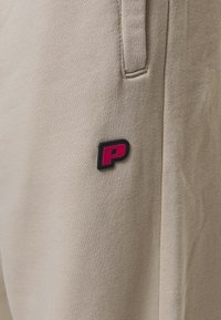 Beige athletic pants feature a textured fabric with a small black and pink logo. Design includes side pockets and relaxed fit.