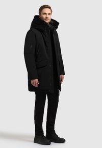 Sort parka med høj krave, store lommer og lynlåsdetaljer. Bæres sammen med sorte bukser og matchende snørebåndsstøvler.