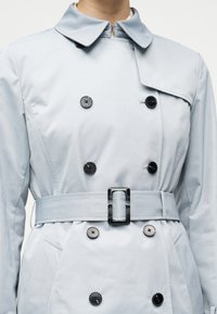 Ανοιχτό γκρι διπλής στήρας καμπαρντίνα με μαύρα και γκρι κουμπιά, ζώνη στη μέση και φαρδύ γιακά σε ένα άτομο, από τους ώμους ως τους γοφούς ορατό.