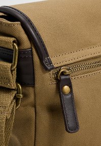 Ruskea canvas-lähettilaukku, jossa tummat nahkakoristeet, kultaiset metalliosat, vetoketjukiinnitys ja teksturoitu kangas. Mukana säädettävä olkahihna ja ompelusaumat.