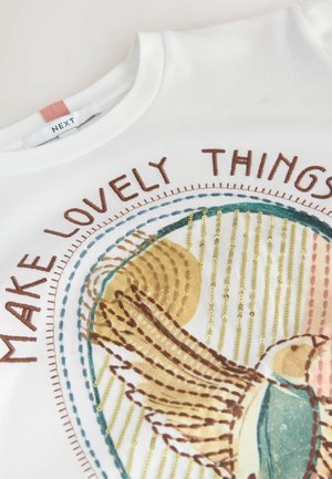 Balta krekls ar izšūtu putna dizainu un frāzi "VEIDOJiet SKAISTAS LIETAS", šūtu brūnā diegā apkārt putna ilustrācijai.