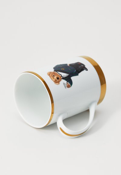 Bijela keramička šalica s zlatnim rubom i naglaskom na dršci, ukrašena medvjedom u odijelu koji drži čašu, leži na boku na bijeloj površini.