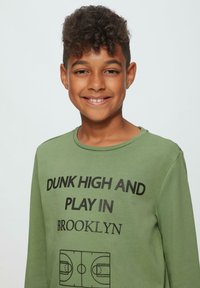 Langärmliger grüner Sweatshirt mit fettem schwarzen Text "DUNK HIGH AND PLAY IN BROOKLYN" und einer Basketballplatzgrafik am unteren Rand.
