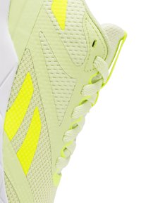Zapatilla deportiva verde claro que presenta materiales de malla y sintéticos, con acentos en amarillo neón, un diseño de cordones intrincado y paneles laterales texturizados.