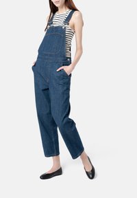 Pantalones de mezclilla con tirantes ajustables, diseño de pierna ancha, bolsillo frontal y puños enrollados, combinados con una camiseta de rayas y zapatos planos negros.