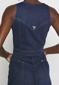 Denimoverall i mörkblått, med en åtsittande bodice, v-formad baksida och silvertriangelns logodetalj. Har platta fickor och kontrasterande sömmar.