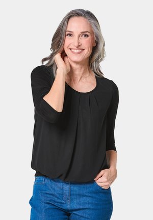 Schwarze Bluse mit großzügigem Ausschnitt, 3/4-Ärmeln und Falten an der Vorderseite. getragen mit blauen Jeans; glattes Material und lockere Passform.