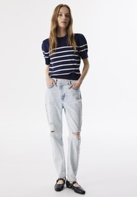 Marine gestreepte trui met korte mouwen, lichtblauwe distressed jeans en zwarte flats met studs, rechtop geplaatst tegen een witte achtergrond.