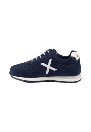 Zapatilla deportiva azul marino con suela blanca, cordones blancos, parte superior de malla y ante, y un gran logo blanco en forma de X en el lateral.