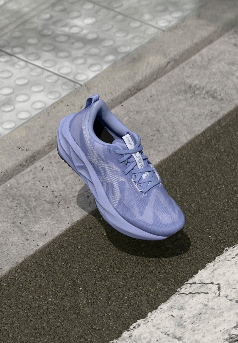 Lyselilla sportssko med teksturert overdel, snøring og polstret såle, plassert på en betongoverflate nær et fotgjengerovergang.