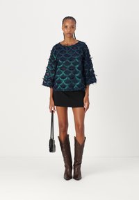 Haut texturé bleu marine avec un motif écaillé, manches évasées, associé à une mini-jupe noire et des bottes de cowboy marron. Sac à main noir à la main.