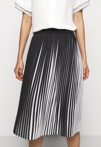 Femme portant une jupe midi plissée à rayures verticales noires et blanches avec une ceinture noire et un chemisier blanc à manches courtes.