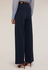 Pantaloni a gamba larga blu navy con tasche laterali, tessuto liscio e vita alta. Abbinati a una camicia a maniche lunghe azzurra e tacchi alti.
