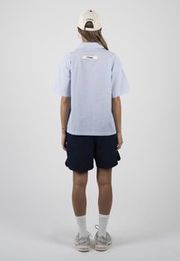 Personne de dos portant une casquette beige, une chemise à manches courtes rayée bleu clair, un short bleu marine, des chaussettes blanches et des baskets grises sur fond gris.