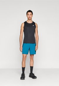 Μαύρο αμάνικο τοπ με στρογγυλή λαιμόκοψη, συνδυασμένο με τιρκουάζ σορτς. Μαύρα αθλητικά παπούτσια με σκούρες κάλτσες, σε ουδέτερο φόντο.