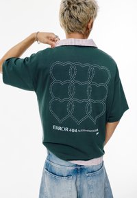 Forest green oversized T-shirt with a checkered collar, featuring an embroidered heart pattern and "ERROR 404 ALTERNATIVE CULT" text.