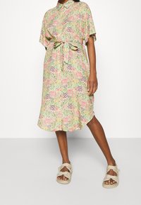 Vestido de camisa floral en colores pastel con cuello, mangas cortas, cintura ceñida con cinturón y dobladillo a la altura de la rodilla. Llevado con sandalias beige.