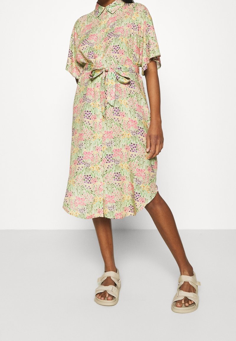 Vestido de camisa floral en colores pastel con cuello, mangas cortas, cintura ceñida con cinturón y dobladillo a la altura de la rodilla. Llevado con sandalias beige.