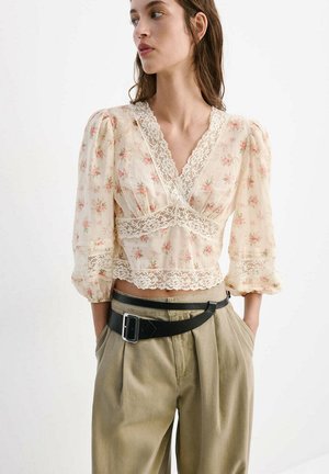 Joven mujer con una blusa de encaje floral color crema con mangas abullonadas y pantalones holgados caqui con cinturón negro, con las manos en los bolsillos, mirando hacia un lado.