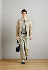 Μπεζ καπιτονέ μπότλερ, ριγέ πουκάμισο, ανοιχτό πράσινες φαρδιές παντελόνες, μαύρη ζώνη, μαύρη τσάντα ώμου, μαύρα παπούτσια, στέκεται σε ξύλινο δάπεδο.