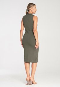 Oliwkowa sukienka bodycon z wysokim dekoltem, bez rękawów, do kolan, z subtelnym detalem wzdłuż tylnego szwu. Noszona z przezroczystymi sandałami na obcasach.