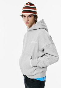 Grijze katoenen hoodie met een kangoeroezak en een logo aan de voorkant. Draagt een blauwe T-shirt eronder en is aangevuld met een gestreepte beanie.