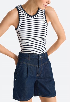 Femme portant un haut blanc sans manches à rayures horizontales noires et un short en jean bleu foncé taille haute avec poches avant et fermeture à boutons.