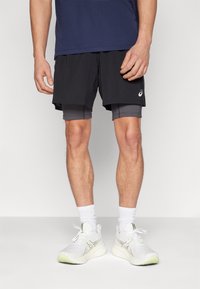Schwarze Sportshorts mit einer inneren grauen Schicht, kombiniert mit weißen Turnschuhen und knöchellangen weißen Socken. Auffälliges Markenlogo auf den Shorts.