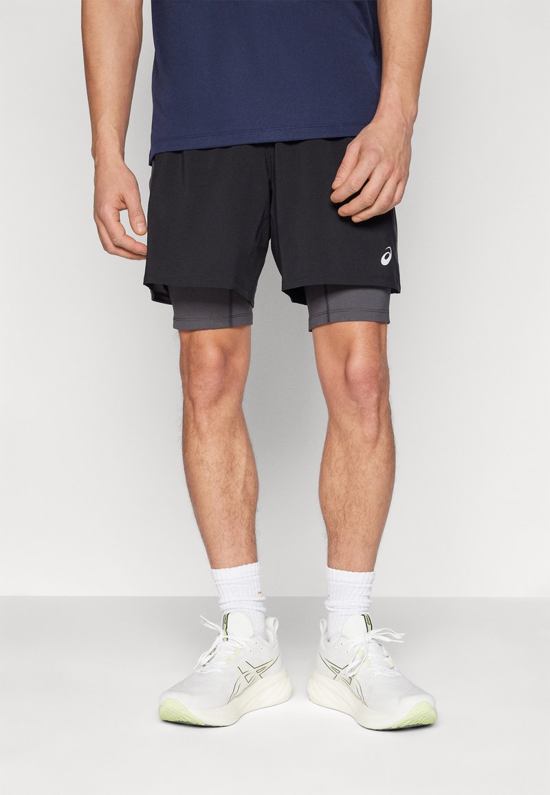 Schwarze Sportshorts mit einer inneren grauen Schicht, kombiniert mit weißen Turnschuhen und knöchellangen weißen Socken. Auffälliges Markenlogo auf den Shorts.