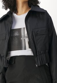 Černá zkrácená bunda z hladké látky se zipovým zapínáním a dvěma předními kapsami, vrstvená přes bílé tričko s grafikou obsahující monochromatický potisk.