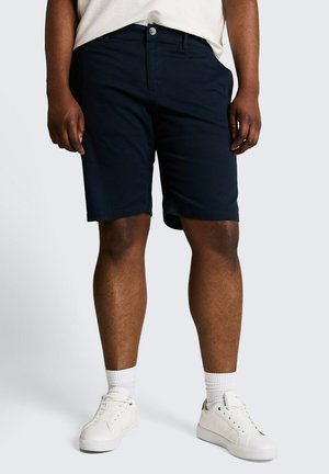 Mężczyzna ubrany w granatowe szorty, białe skarpetki typu crew i białe tenisówki, stojący na jasnoszarym tle.