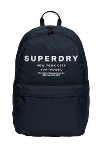Granatowy plecak z tkaniny o zaokrąglonym kształcie, z białym nadrukowanym logo "SUPERDRY" oraz przednią kieszenią na zamek do przechowywania.
