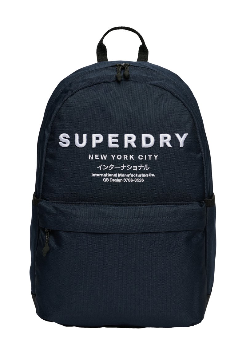 Granatowy plecak z tkaniny o zaokrąglonym kształcie, z białym nadrukowanym logo "SUPERDRY" oraz przednią kieszenią na zamek do przechowywania.