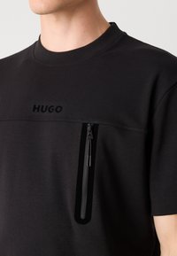 Svart kortermet t-skjorte med rund hals, med en frontlomme med glidelås og preget logo på brystet.