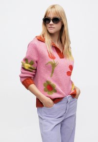 Ροζ πλεκτή μπλούζα με λουλουδένια σχέδια σε πράσινο, κόκκινο και κίτρινο; πορτοκαλί γιακάς; συνδυασμένη με ανοιχτό μωβ παντελόνι από κοτλέ.