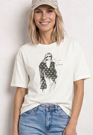 Lächelnde Frau mit blonden Haaren, die eine gemusterte Baseballkappe, ein weißes T-Shirt mit schwarzer Polka-Dot-Modeillustration und hellblaue Jeans trägt.