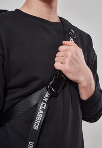 Sort crossbody-strop med justerbart spænde, prydet med vævet tekst "URBAN CLASSICS." Glat tekstur, præsenterer et minimalistisk design.