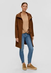 Brauner Wollmantel mit einem gerippten Kragen, beigem Oberteil, blauen Skinny-Jeans und beigen Sneakers. Der Mantel hat eine Knopfleiste vorne und ein langes Design.