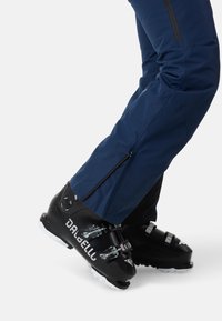 Svarta skidpjäxor med metallspännen, ihopparat med blå skidbyxor med sidodragkedjor. Synlig fotpositionering på en vit bakgrund.